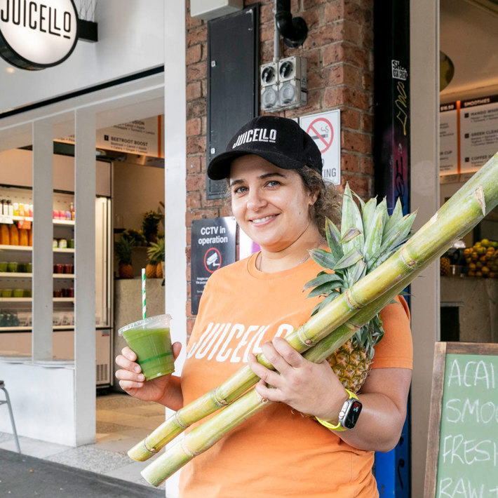 Person in an orange t-shirt, holding a green smoothie and a large stalk of sugar cane with a pineapple attached, standing in front of ‘JUICELO’, a juice bar.